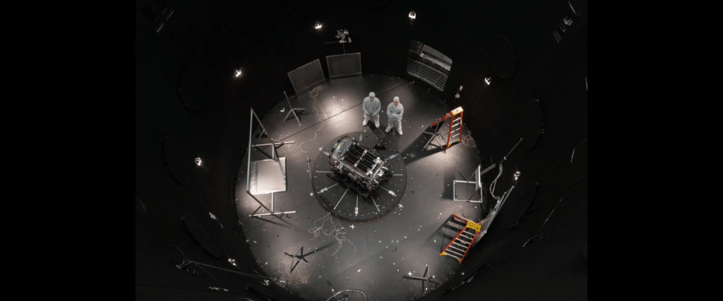 Astronauts or engineers in white suits stand over a central experimental module in a circular, dimly lit test chamber filled with cables and scaffolding pieces.