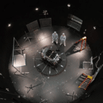 Astronauts or engineers in white suits stand over a central experimental module in a circular, dimly lit test chamber filled with cables and scaffolding pieces.