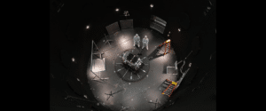 Astronauts or engineers in white suits stand over a central experimental module in a circular, dimly lit test chamber filled with cables and scaffolding pieces.
