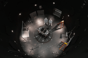 Astronauts or engineers in white suits stand over a central experimental module in a circular, dimly lit test chamber filled with cables and scaffolding pieces.