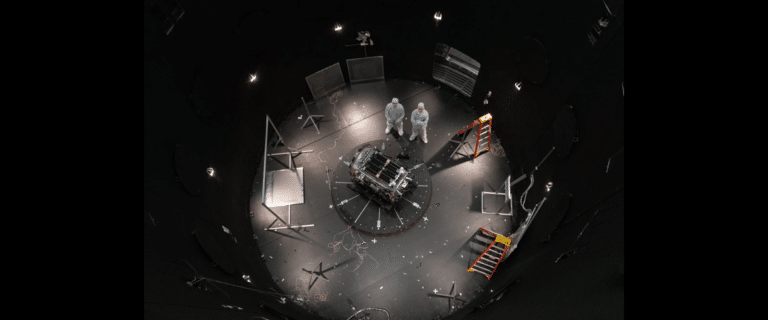 Astronauts or engineers in white suits stand over a central experimental module in a circular, dimly lit test chamber filled with cables and scaffolding pieces.