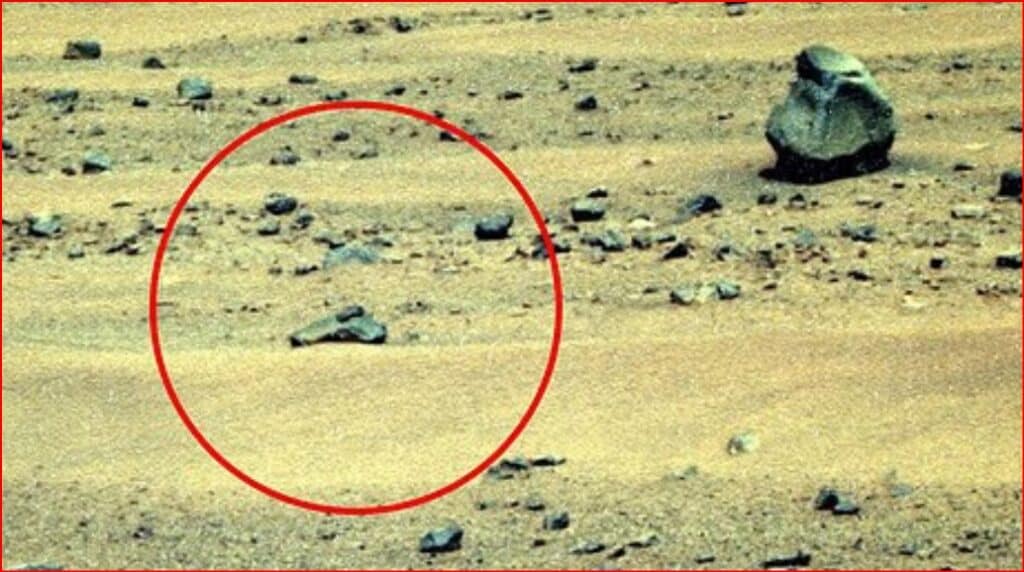 A barren rocky landscape with a circled rock formation highlighted in red in the foreground, suggesting a point of interest on the ground surface on a dusty terrain.