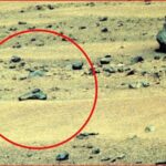 A barren rocky landscape with a circled rock formation highlighted in red in the foreground, suggesting a point of interest on the ground surface on a dusty terrain.