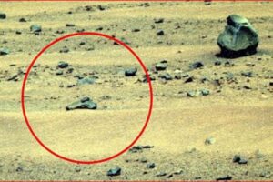 A barren rocky landscape with a circled rock formation highlighted in red in the foreground, suggesting a point of interest on the ground surface on a dusty terrain.