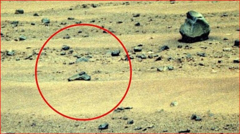 A barren rocky landscape with a circled rock formation highlighted in red in the foreground, suggesting a point of interest on the ground surface on a dusty terrain.