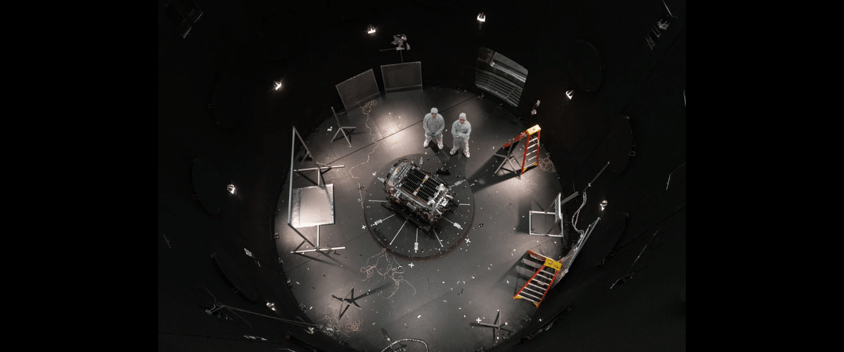 Astronauts or engineers in white suits stand over a central experimental module in a circular, dimly lit test chamber filled with cables and scaffolding pieces.