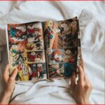 Person's hands hold open a colorful comic book laid on white sheets, panels show superheroes in action.