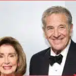 Smiling elderly couple dressed for a formal event; woman in a light-colored outfit with earrings, man in a black tuxedo and bow tie.