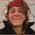Woman with a red headband and tousled hair smiling at the camera inside a tent, wearing a black jacket.