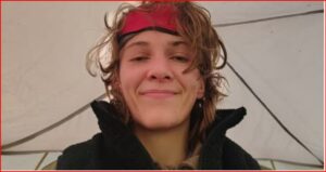 Woman with a red headband and tousled hair smiling at the camera inside a tent, wearing a black jacket.