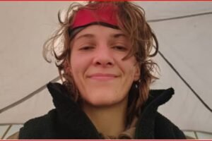 Woman with a red headband and tousled hair smiling at the camera inside a tent, wearing a black jacket.