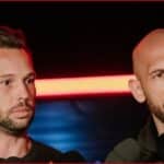 Two men at a press conference; one speaks into microphones while the other listens nearby.