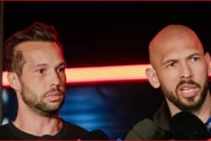 Two men at a press conference; one speaks into microphones while the other listens nearby.