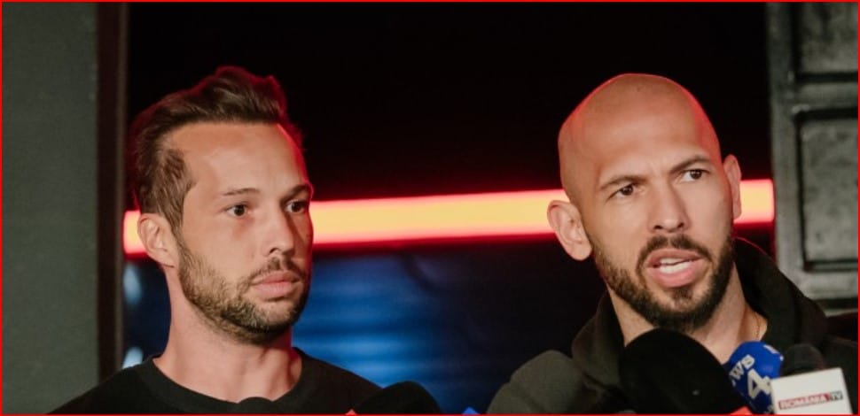 Two men at a press conference; one speaks into microphones while the other listens nearby.