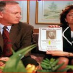 Man and woman in business attire sit on a couch, holding a $100,000 reward poster featuring a woman's photo in a waiting-room setting.