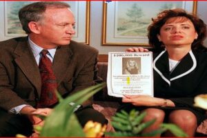 Man and woman in business attire sit on a couch, holding a $100,000 reward poster featuring a woman's photo in a waiting-room setting.