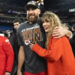 A smiling man in a Kansas City Chiefs t‑shirt and cap stands with a woman in an orange sweater hugging him on a football field during a victory celebration, with photographers nearby.