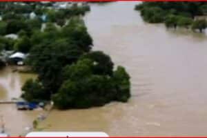 Drone view of a residential neighborhood flooded, with brown water covering streets, yards, and driveways around trees and houses partially submerged.