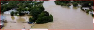Drone view of a residential neighborhood flooded, with brown water covering streets, yards, and driveways around trees and houses partially submerged.