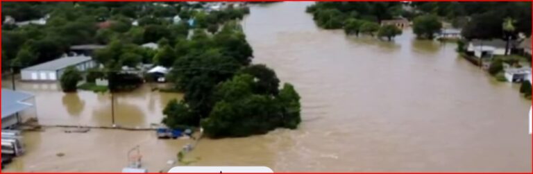 Drone view of a residential neighborhood flooded, with brown water covering streets, yards, and driveways around trees and houses partially submerged.