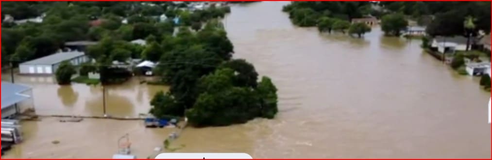 Drone view of a residential neighborhood flooded, with brown water covering streets, yards, and driveways around trees and houses partially submerged.