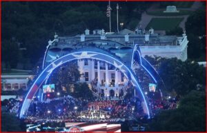Nighttime view of the White House lawn with blue-lit arches, a large crowd, and a concert taking place on stage.