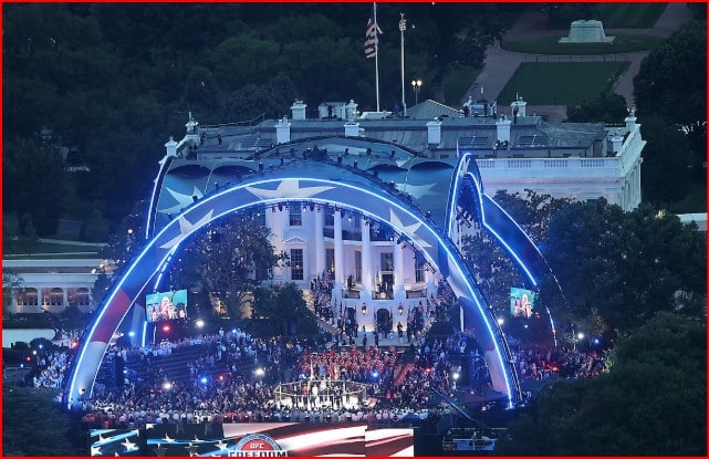 Nighttime view of the White House lawn with blue-lit arches, a large crowd, and a concert taking place on stage.