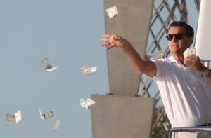 Man wearing sunglasses in a white shirt throws dollar bills into the air against a blue sky.