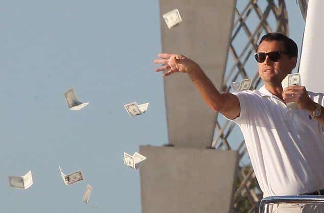 Man wearing sunglasses in a white shirt throws dollar bills into the air against a blue sky.