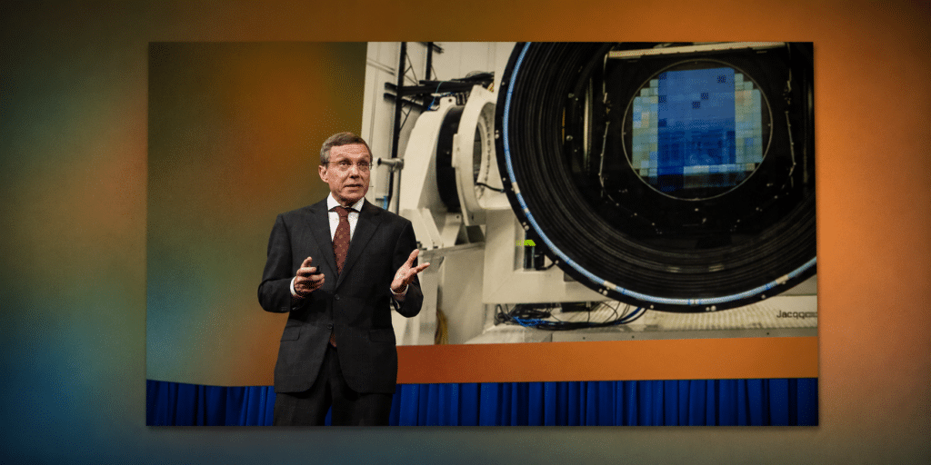 Man in a dark suit giving a presentation beside a large scientific device with a circular opening and a blue heat-shield, on a stage with orange walls and blue curtains behind him