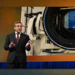 Man in a dark suit giving a presentation beside a large scientific device with a circular opening and a blue heat-shield, on a stage with orange walls and blue curtains behind him