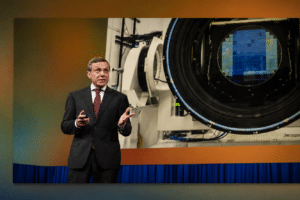 Man in a dark suit giving a presentation beside a large scientific device with a circular opening and a blue heat-shield, on a stage with orange walls and blue curtains behind him