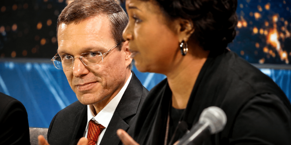 Man in a suit with red tie looks toward camera while a woman speaks into a microphone beside him on a panel set.