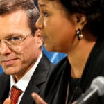 Man in a suit with red tie looks toward camera while a woman speaks into a microphone beside him on a panel set.