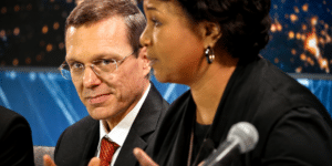 Man in a suit with red tie looks toward camera while a woman speaks into a microphone beside him on a panel set.