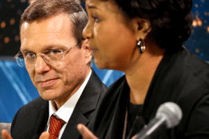 Man in a suit with red tie looks toward camera while a woman speaks into a microphone beside him on a panel set.