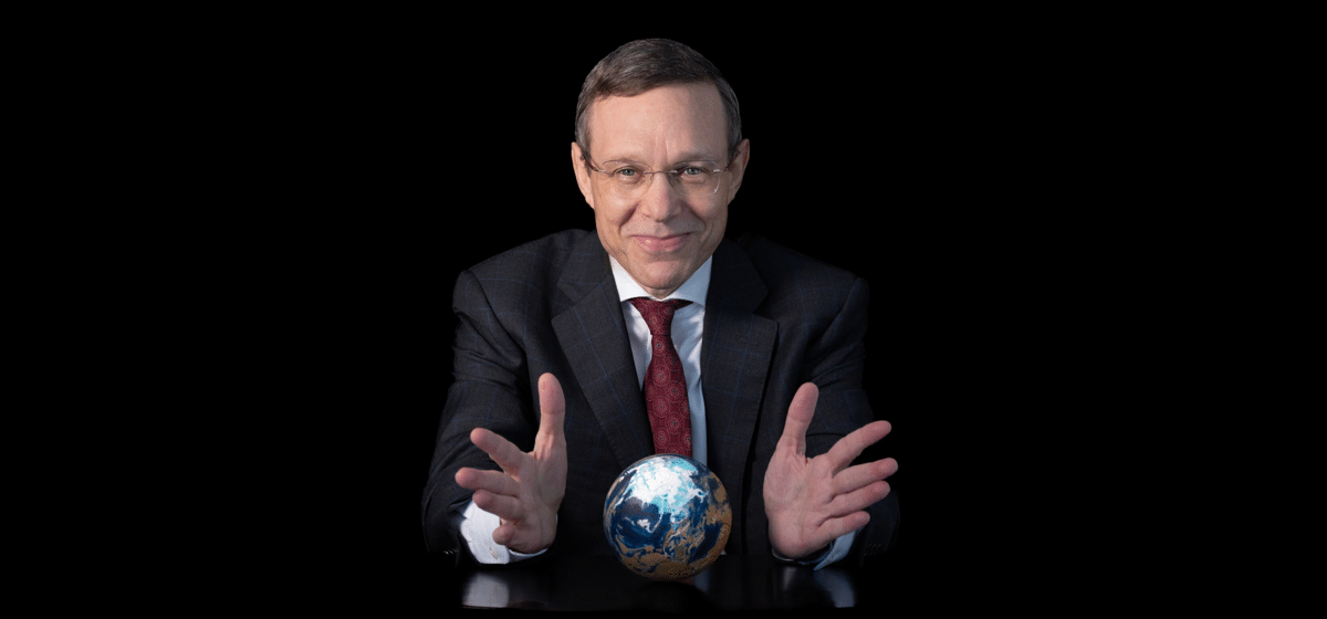 Smiling businessman in a dark suit holds a small globe on a table, appearing to present the Earth against a black background.