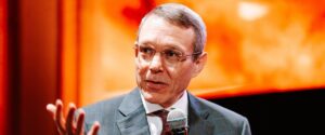 Man in a gray pinstripe suit speaks into a microphone on stage, gesturing with his hand against an orange-lit backdrop.