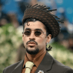 Man in a brown suit, beige tie, and round sunglasses wearing a woven brown hat at a formal event with a blurred crowd behind him.