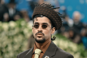 Man in a brown suit, beige tie, and round sunglasses wearing a woven brown hat at a formal event with a blurred crowd behind him.