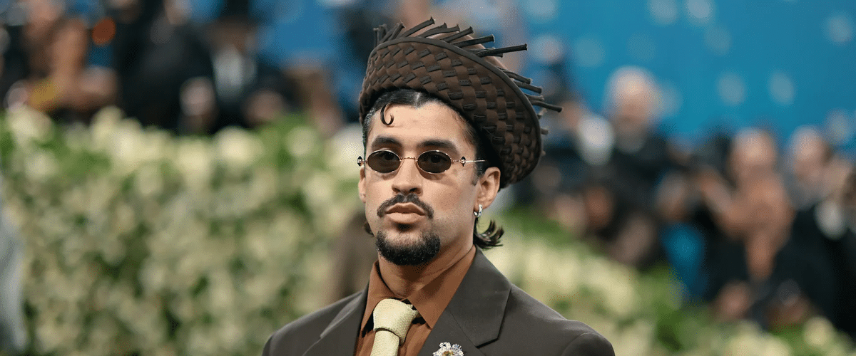 Man in a brown suit, beige tie, and round sunglasses wearing a woven brown hat at a formal event with a blurred crowd behind him.