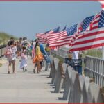 Pedestrians stroll along a sunny boardwalk lined with waving American flags along the railing.
