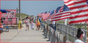 Pedestrians stroll along a sunny boardwalk lined with waving American flags along the railing.