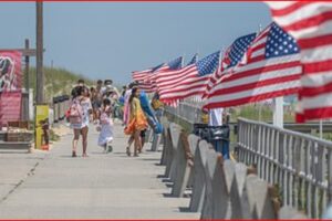 Pedestrians stroll along a sunny boardwalk lined with waving American flags along the railing.
