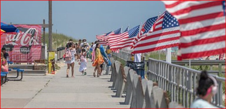 Pedestrians stroll along a sunny boardwalk lined with waving American flags along the railing.