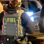 Group of FBI agents in tactical vests and gear around a vehicle at night, one vest clearly shows 'FBI' on the back