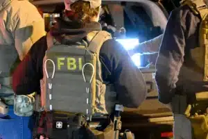 Group of FBI agents in tactical vests and gear around a vehicle at night, one vest clearly shows 'FBI' on the back