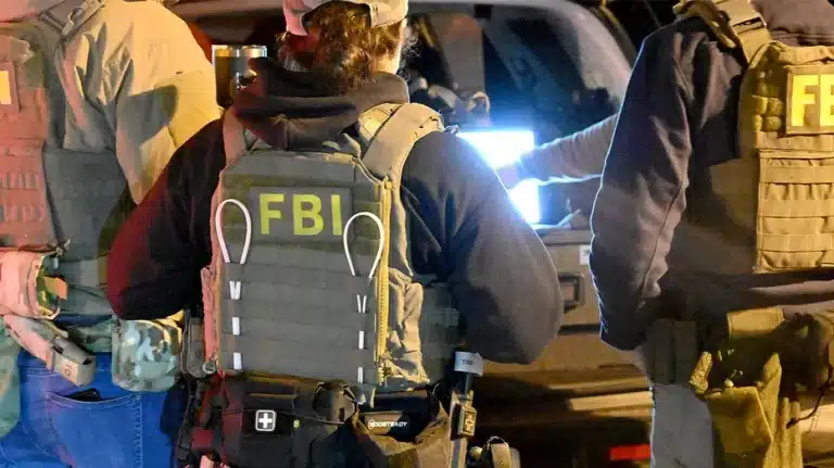 Group of FBI agents in tactical vests and gear around a vehicle at night, one vest clearly shows 'FBI' on the back