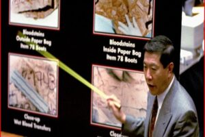 Man in a suit points to a forensic display showing bloodstain evidence in paper bags on a dark board.