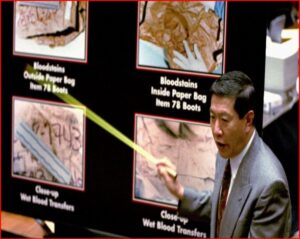 Man in a suit points to a forensic display showing bloodstain evidence in paper bags on a dark board.